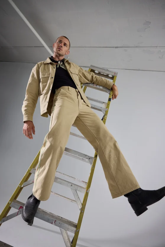Contemporary menswear editorial showing model in mid-motion wearing beige clothing on light background.