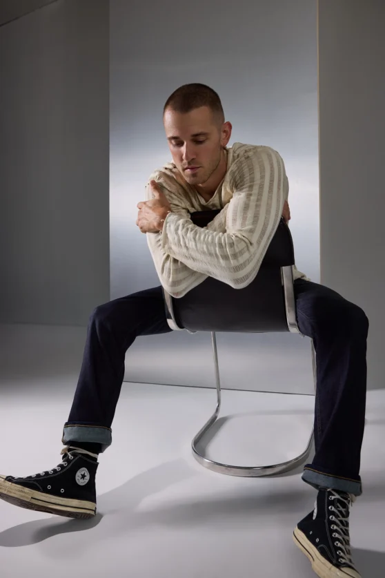 Editorial fashion shot of a male model in knitwear, seated on a transparent chair for ecommerce lookbook.
