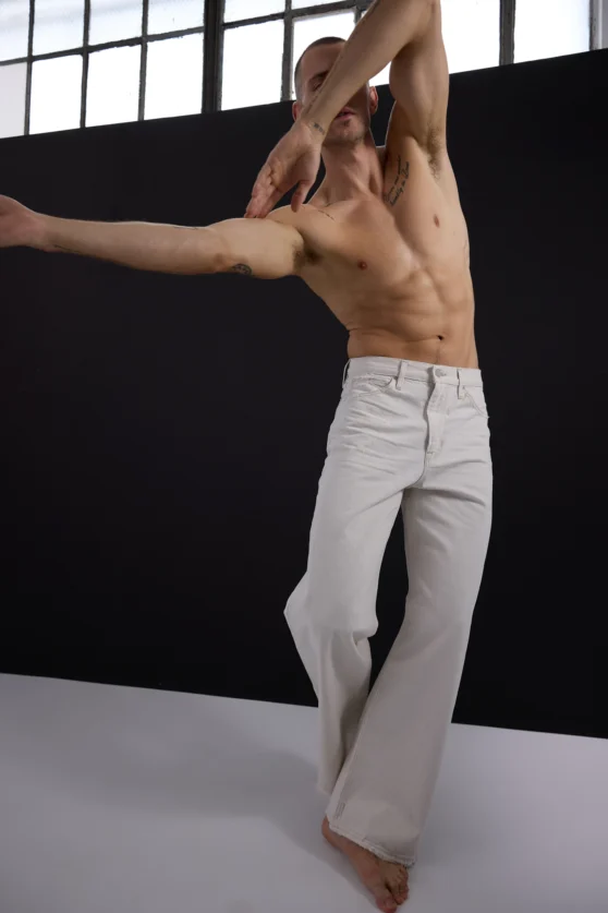 Minimalist fashion photo of shirtless male model stretching in white pants against a black background.