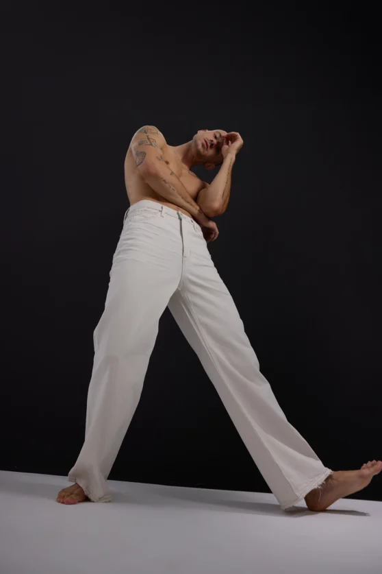 Shirtless male model in white trousers performing strong fashion stance on black backdrop for ecommerce styling.