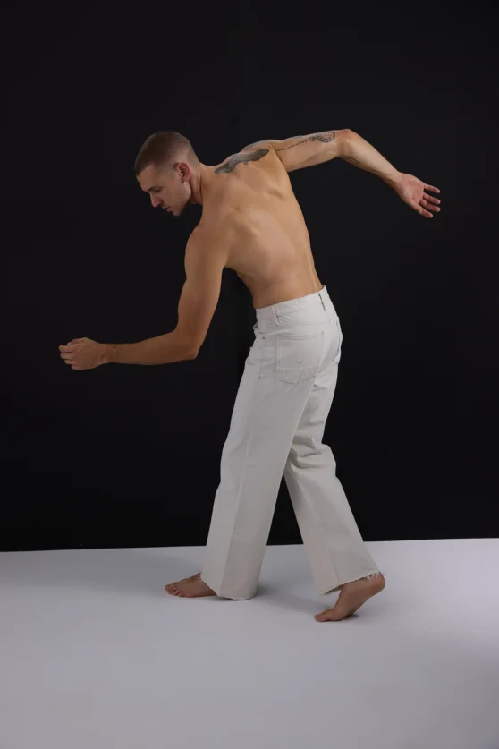 Tattooed male model in white pants striking expressive editorial pose in high-fashion studio shoot.